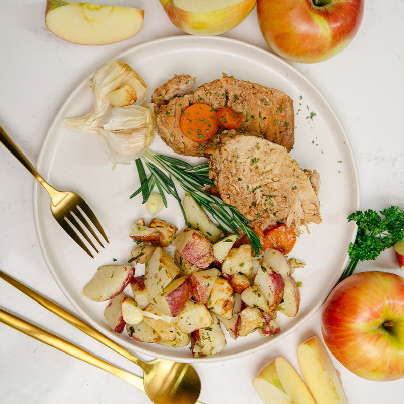 Plated dish with sliced Beehive Meals Apple Cider Pork Roast with a side of carrots and roasted red potatoes and apples surrounding the plate on a white surface.