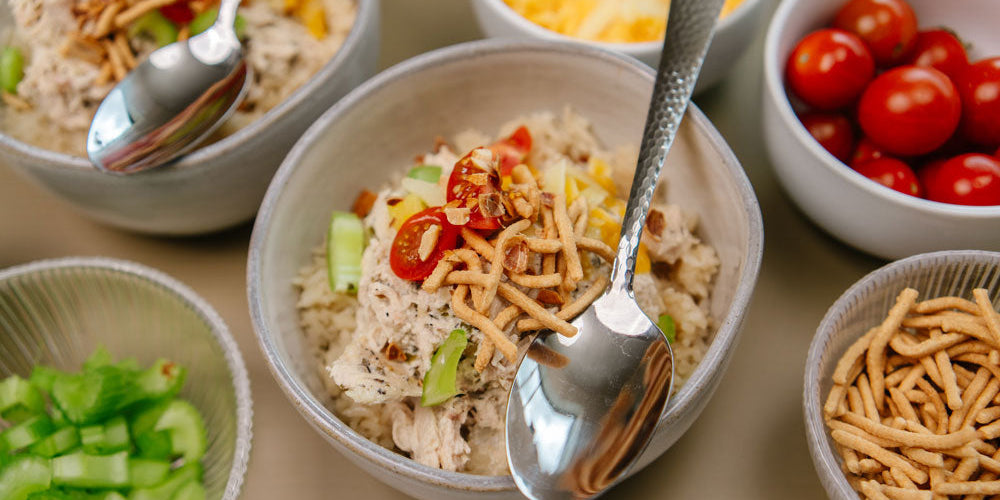 Gray bowl with shredded chicken, crunch noodles, tomatoes and celery with a silver spoon.