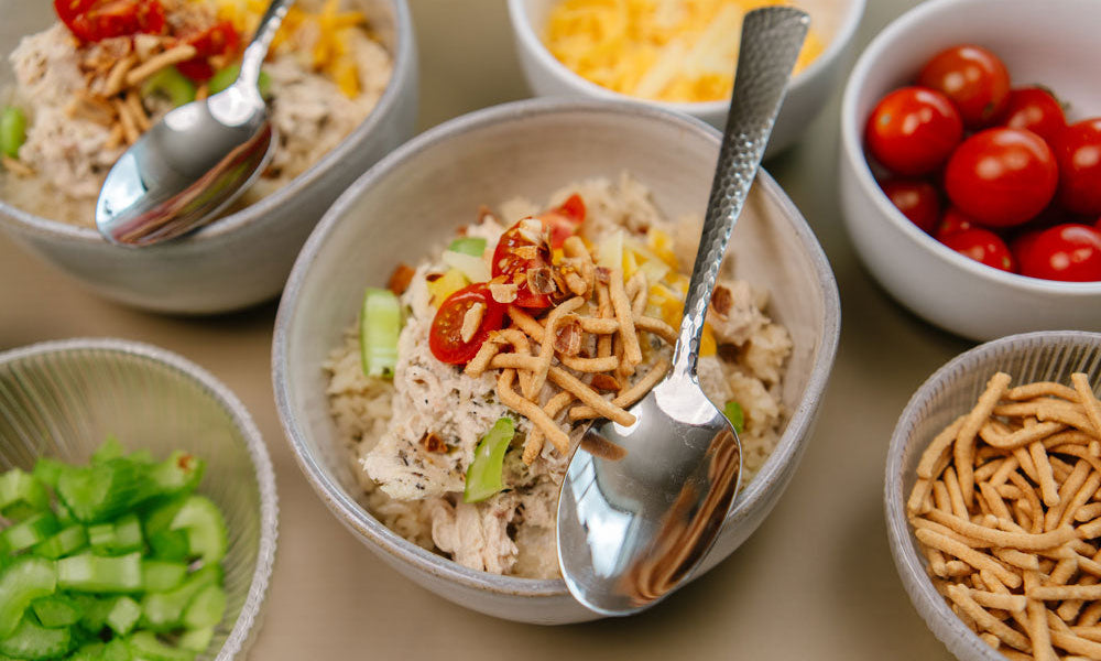Gray bowl with shredded chicken, crunch noodles, tomatoes and celery with a silver spoon.