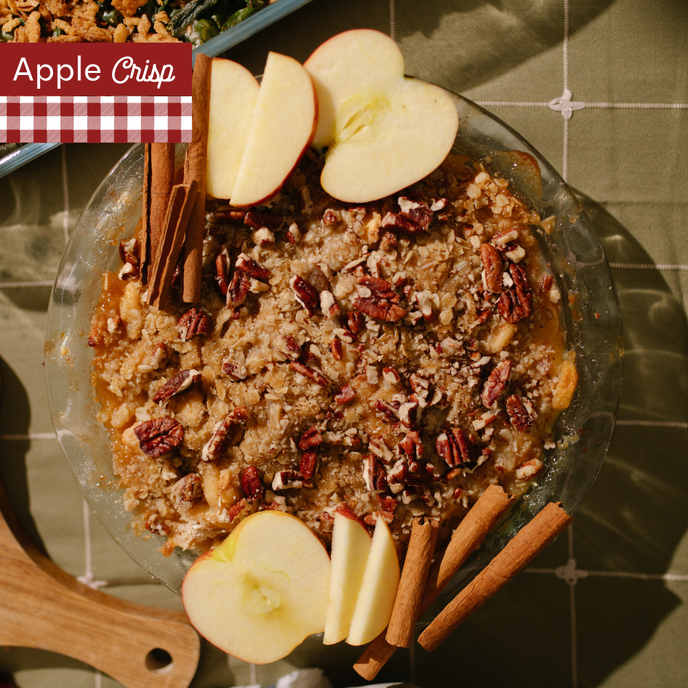 Apple Crisp with a crumble topping in a glass pie dish with fresh apple halves and cinnamon sticks next to the dish. A red-and-white gingham banner at the top left reads 'Apple Crisp'.