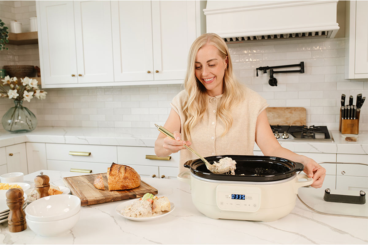 Allyse Jackson, Founder, serving a Beehive Meal using a cream-colored slow cooker with a black insert in a bright, modern kitchen. She is holding a spoon with food inside the pot.