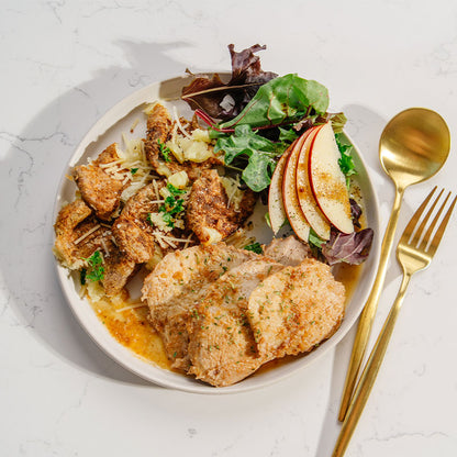 Plated dish of sliced pork with potatoes, a salad and apples, served on a light surface with gold cutlery.