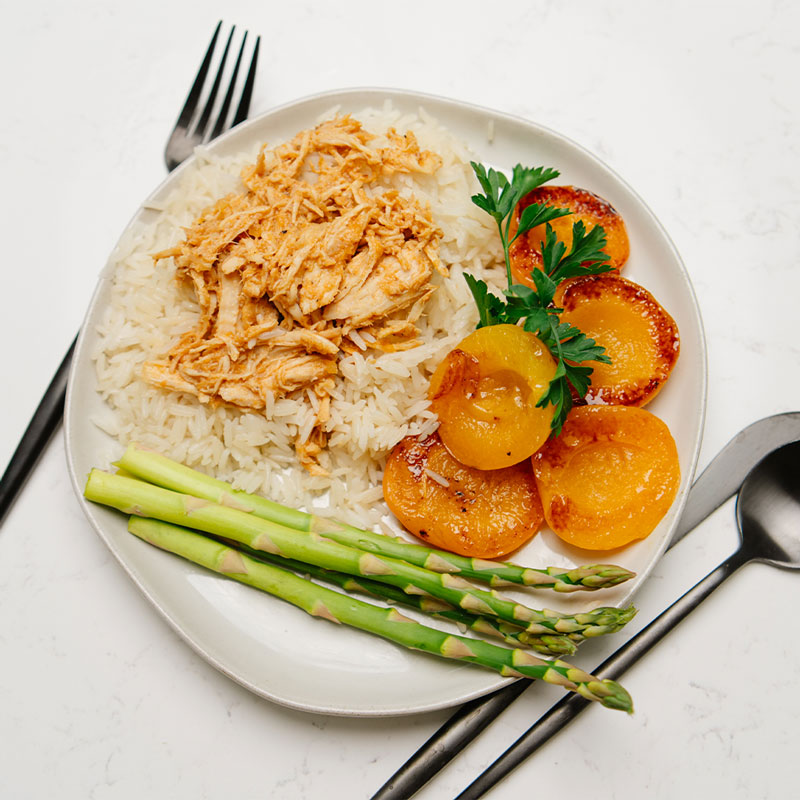 Beehive Meals Apricot Chicken shredded with rice, chicken, apricots, and asparagus on a white background with a black fork, knife and spoon.