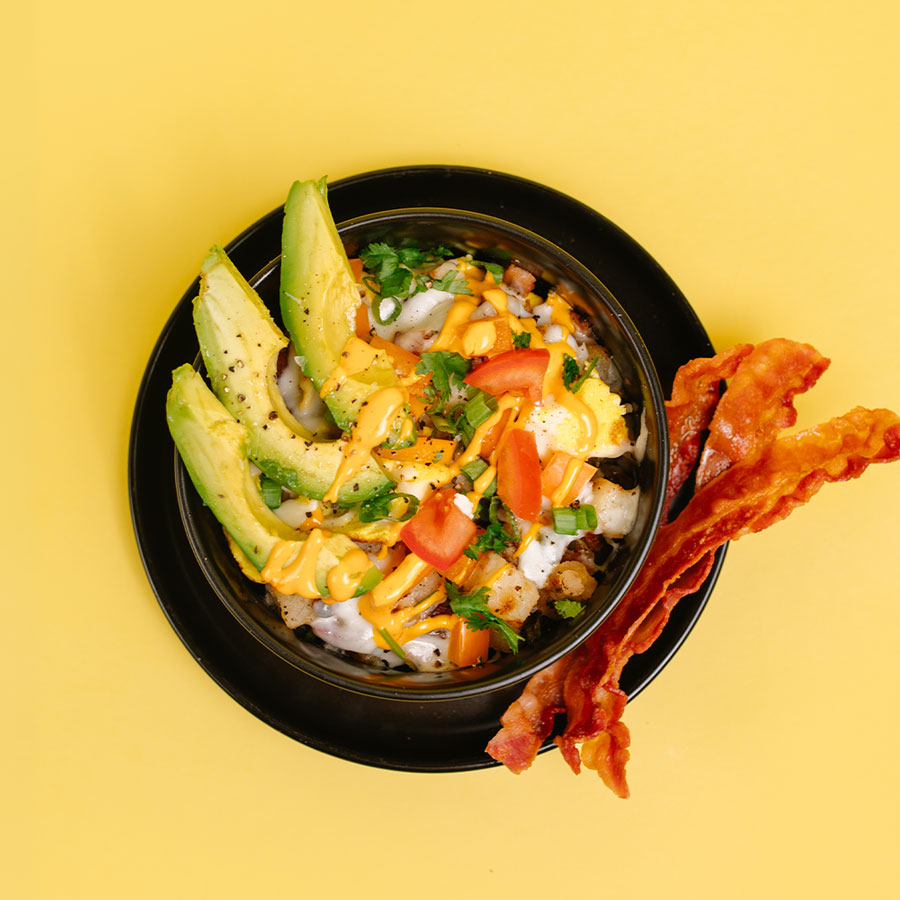 A top-down view of a loaded breakfast bowl on a black plate, set against a solid yellow background. The bowl contains a mixture of eggs, cheese, and bacon, topped with avocado slices, diced tomatoes, and chopped parsley.