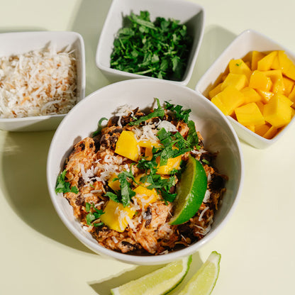Bowl of Beehive Meals Chicken Burrito Bowls with mango and lime, surrounded by smaller bowls of ingredients on a light background