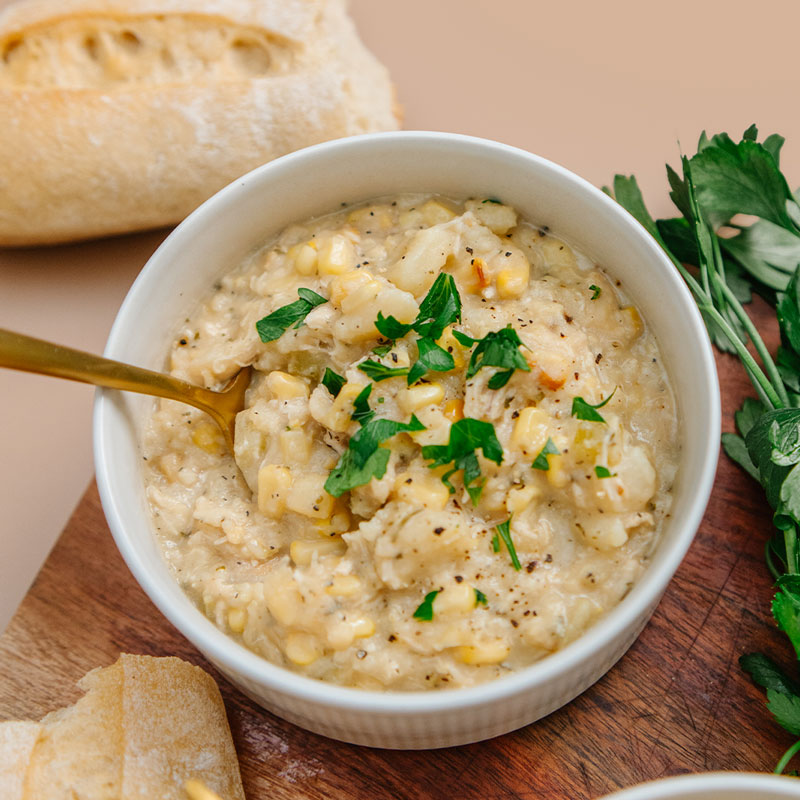 White bowl of creamy corn and chicken soup with a gold spoon, surrounded by bread and herbs on a wooden board.