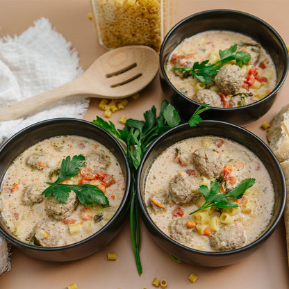 Three bowls of meatball soup with pasta and vegetables on a wooden surface.