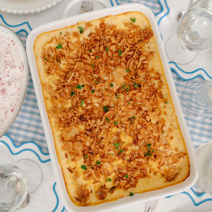 Rectangular dish of cheesy potatoes with french fried onions on top on a tablecloth with wine glasses.