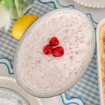 Dessert with raspberries in a bowl, placed on a tablecloth with a lemon and vase in the background.