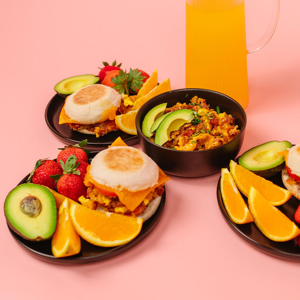 Colorful breakfast setting with sandwiches, bowls, and fruits on a pink background