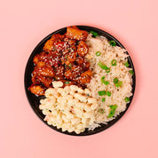 Black plate with pineapple bbq chicken, macaroni salad, and rice on a pink background