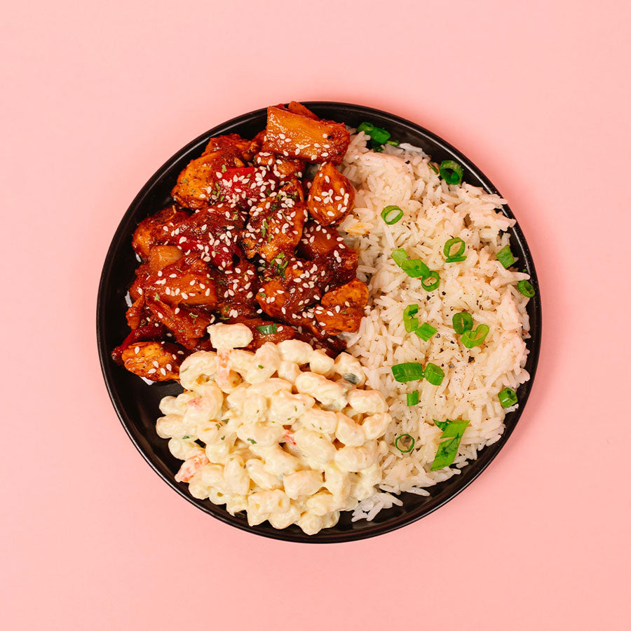 Black plate with pineapple bbq chicken, macaroni salad, and rice on a pink background