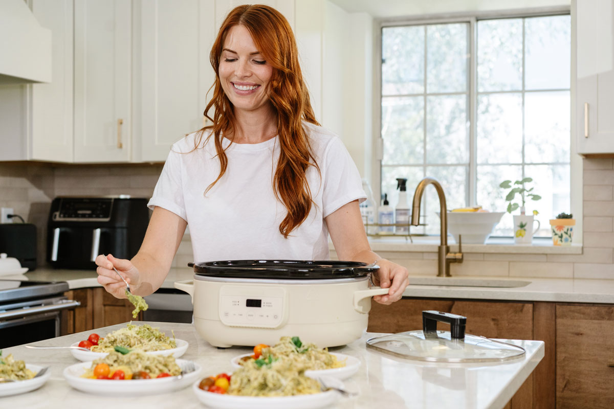 Woman in a kitchen preparing food using a slow cooker placing contents of the slow cooker on a white plate.