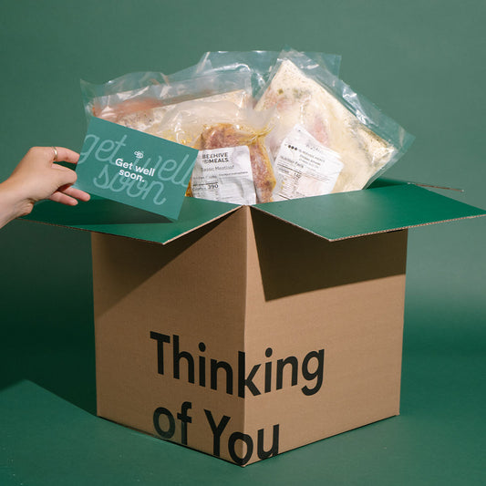A cardboard gift box with the words 'Thinking of You' printed on the front, filled with packaged sandwiches and a teal card that says 'get well soon.' A hand is holding the card.