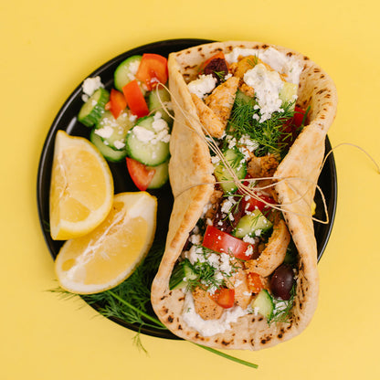A top-down view of a Greek pita sandwich filled with grilled chicken, feta cheese, cucumber, tomato, and dill, served on a black plate with lemon wedges and a side salad on a bright yellow background.
