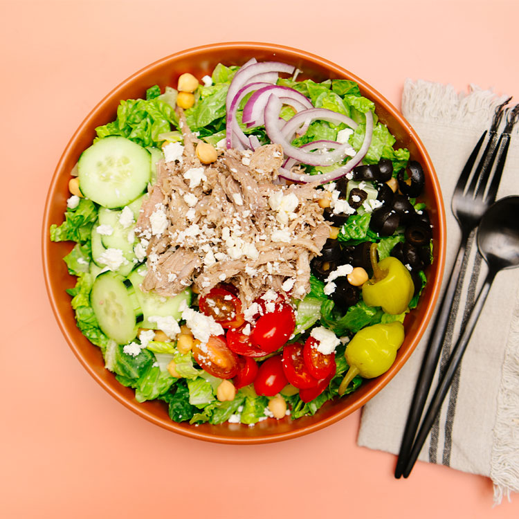 Salad with greens, Beehive Meals Greek Pork, cucumbers, tomatoes, olives, pepperoncinis and chickpeas in a brown bowl with a black fork and spoon on a peach colored background.