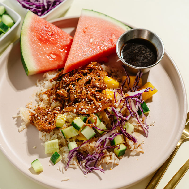Plate of shredded pork with rice, vegetables, and watermelon slices on a white plate.