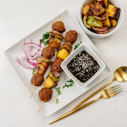Plated Beehive Meals Hawaiian Teriyaki Meatballs skewered with bell peppers and pineapple, and a side of dipping sauce on a white surface.