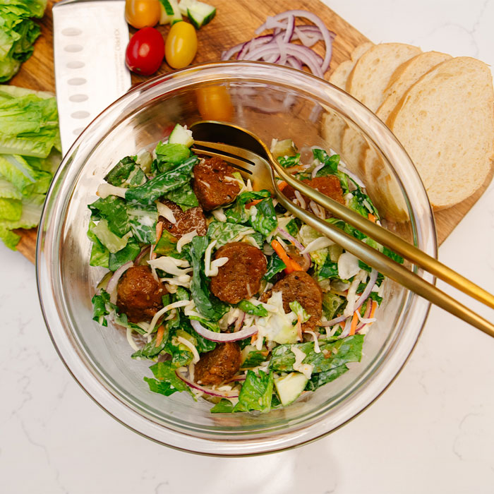 Clear glass bowl with salad mixture and cut up honey buffalo meatballs with a gold spoon and fork.