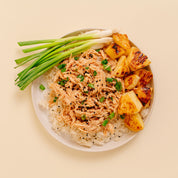 A top-down view of a white plate containing shredded Huli Huli Chicken over white rice, topped with sesame seeds and chopped green onions, served with grilled pineapple chunks on a light beige background.