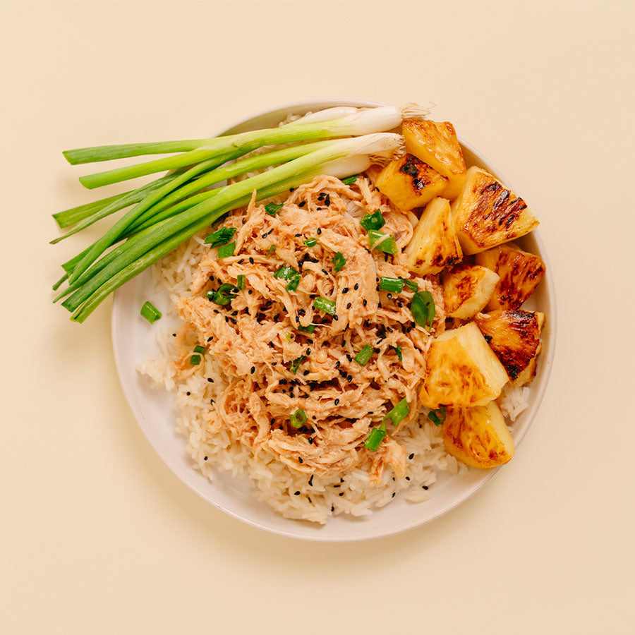 A top-down view of a white plate containing shredded Huli Huli Chicken over white rice, topped with sesame seeds and chopped green onions, served with grilled pineapple chunks on a light beige background.