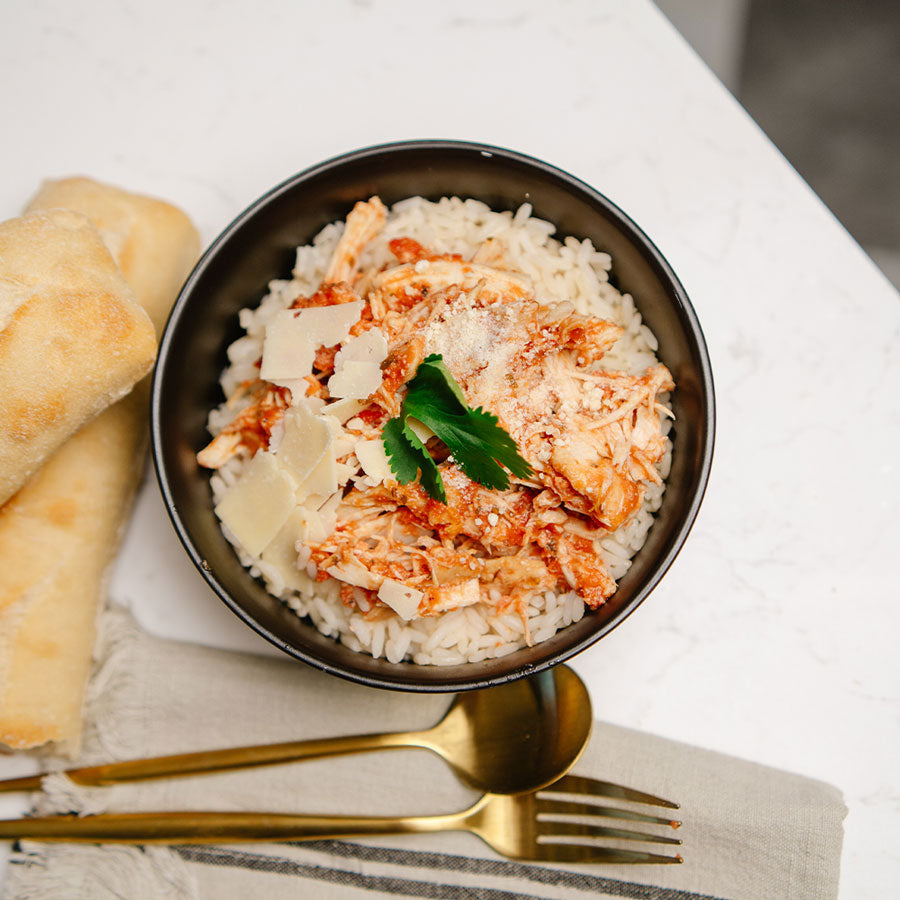 A black bowl with rice topped with Beehive Meals Italian Chicken with bread and a gold fork and spoon on a white background