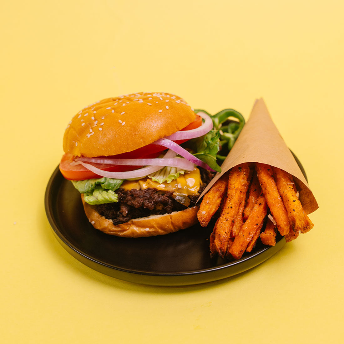 A hamburger with sesame bun, lettuce, tomato, red onion, and melted cheese served on a black plate next to a cone of sweet potato french fries on a solid yellow background.