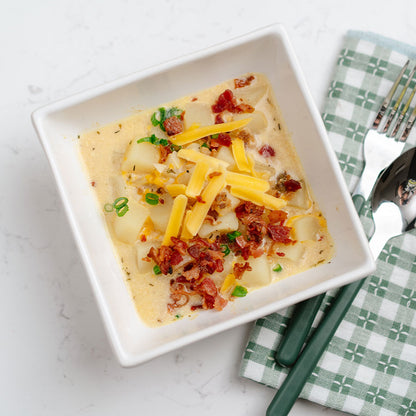 Square white bowl with Beehive Meals Loaded Cheesy Potato Soup on a light surface with checkered napkin and utensils.