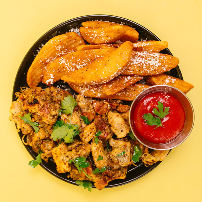 A top-down view of a black plate filled with golden-brown sweet potato wedges dusted with powdered sugar, seasoned chicken and rice, and a small metal cup of ketchup on a solid yellow background.