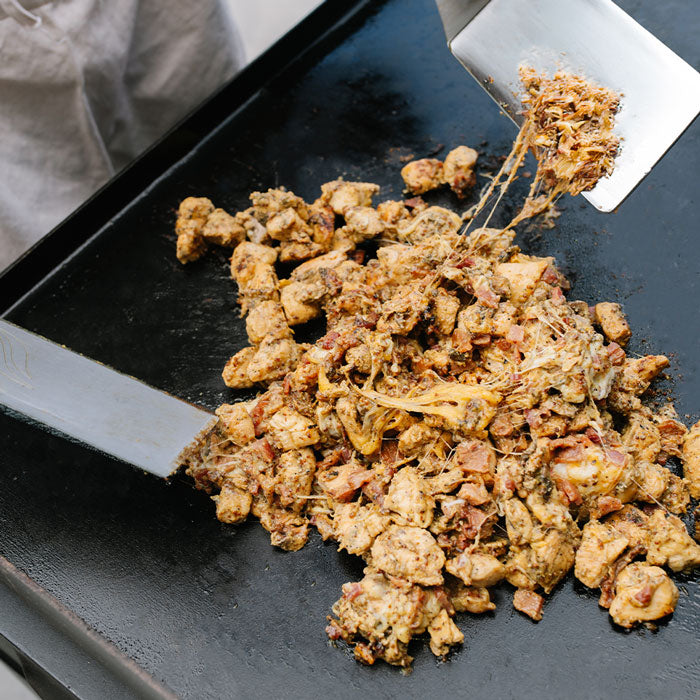 Close-up of cooked food being scraped from a black flat-top grill with metal spatulas, showing a mixture of browned pieces and melted yellow cheese.