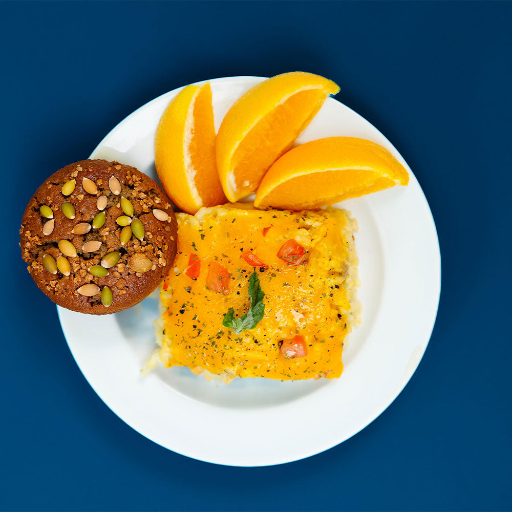 Breakfast casserole on a white plate with orange slices and a muffin against a blue background