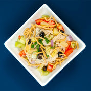 Colorful salad with noodles, vegetables, and olives in a white bowl on a blue background
