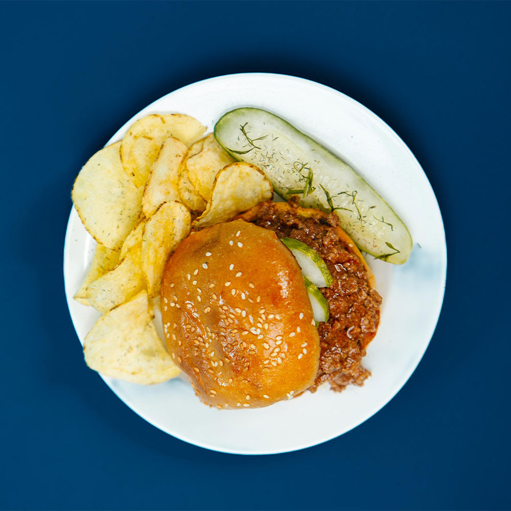 Ground Beef with sesame bun, pickles and chips on a white plate against a blue background