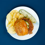 Ground Beef with sesame bun, pickles and chips on a white plate against a blue background