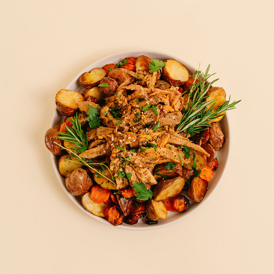 A top-down view of a prepared food dish, featuring shredded cooked meat mixed with roasted potatoes, carrots, and green herbs, served on a light-colored plate against a solid beige background.