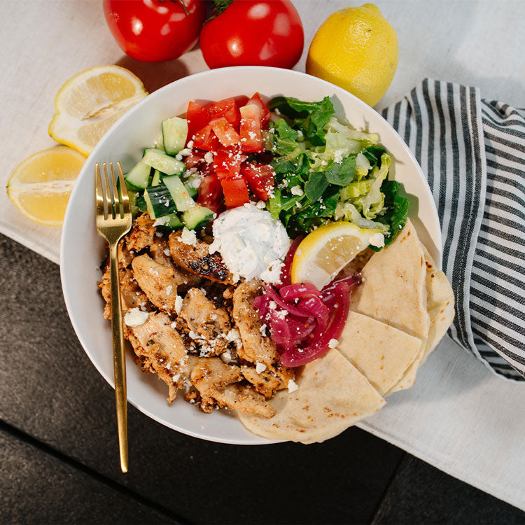 Plated dish with chicken, salad, and pita bread on a table with lemons and tomatoes.