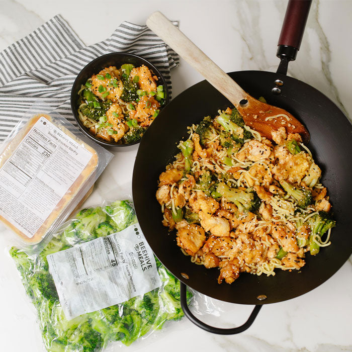 Wok with stir-fried vegetables and chicken on a marble countertop with ingredients and packaging.
