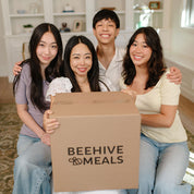Four people holding a box with 'Beehive Meals' logo in a home setting