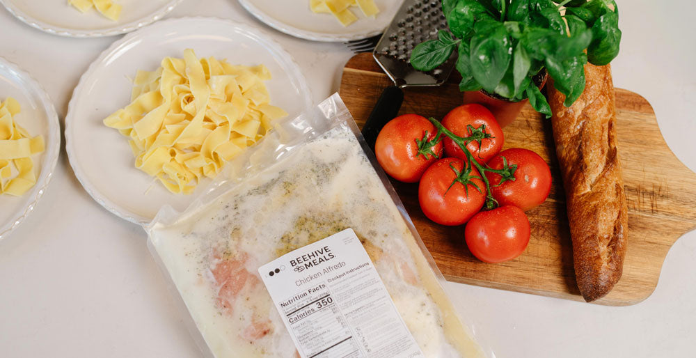 A package of Beehive Meals Chicken Alfredo is shown on a white surface next to plates of prepared pasta, fresh tomatoes, basil, and a baguette on a wooden cutting board.