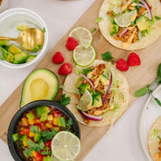 Tacos on a wooden board with avocado, lime, and raspberries