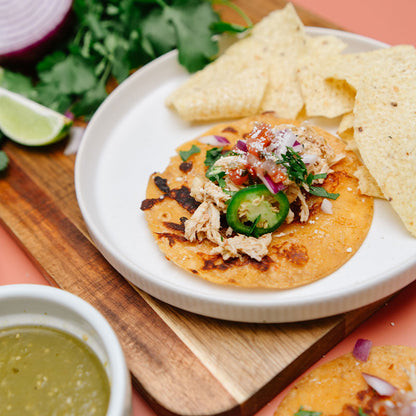 White plate with a corn tortilla laying flat with Beehive Meals Salsa Verde Chicken topped with pico and tortilla chips on the side on a wooden cutting board with a pink background.