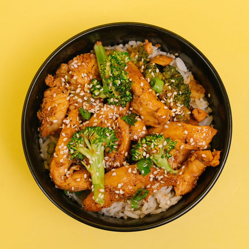 A top-down view of a black bowl filled with cooked chicken, broccoli florets, white sesame seeds, and white rice, set against a solid yellow background.