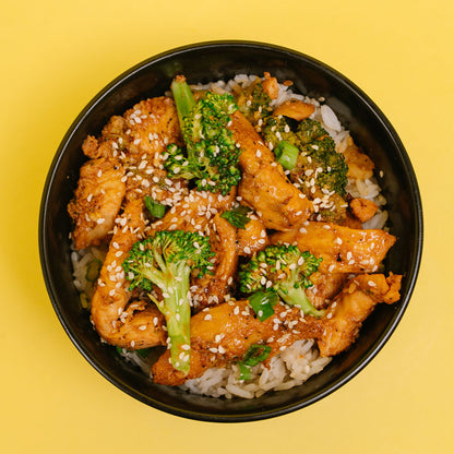A top-down view of a black bowl filled with cooked chicken, broccoli florets, white sesame seeds, and white rice, set against a solid yellow background.