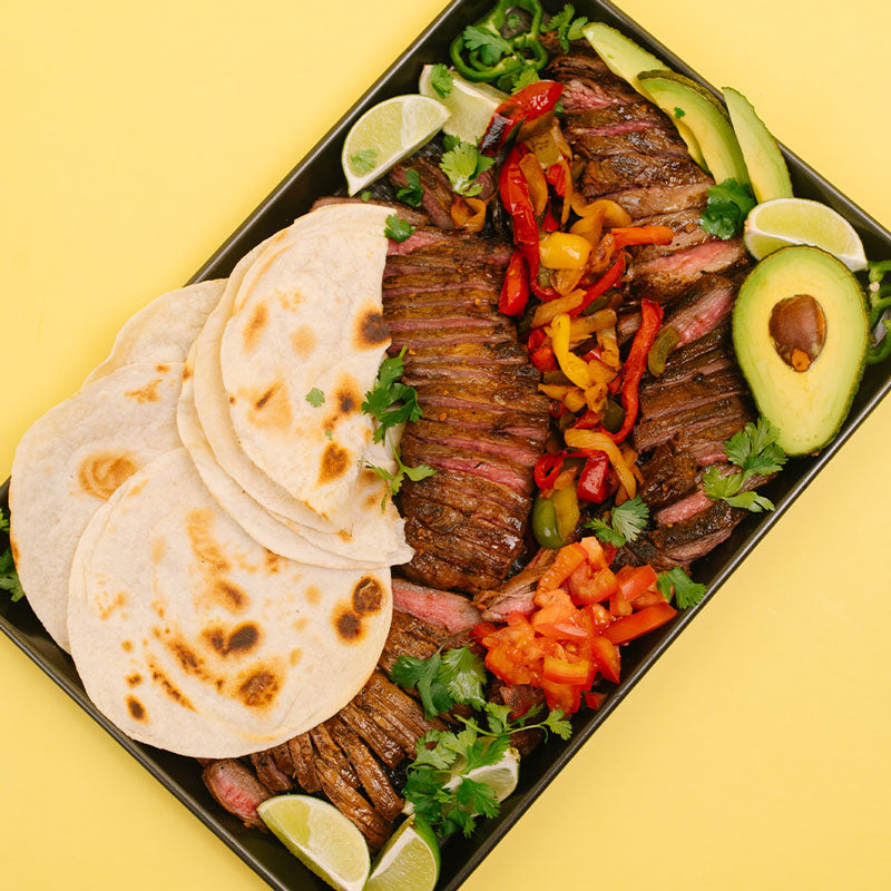 A black rectangular platter filled with grilled sliced meat, colorful bell pepper strips, diced tomatoes, avocado slices, lime wedges, and stacked tortillas, set against a solid pale yellow background.