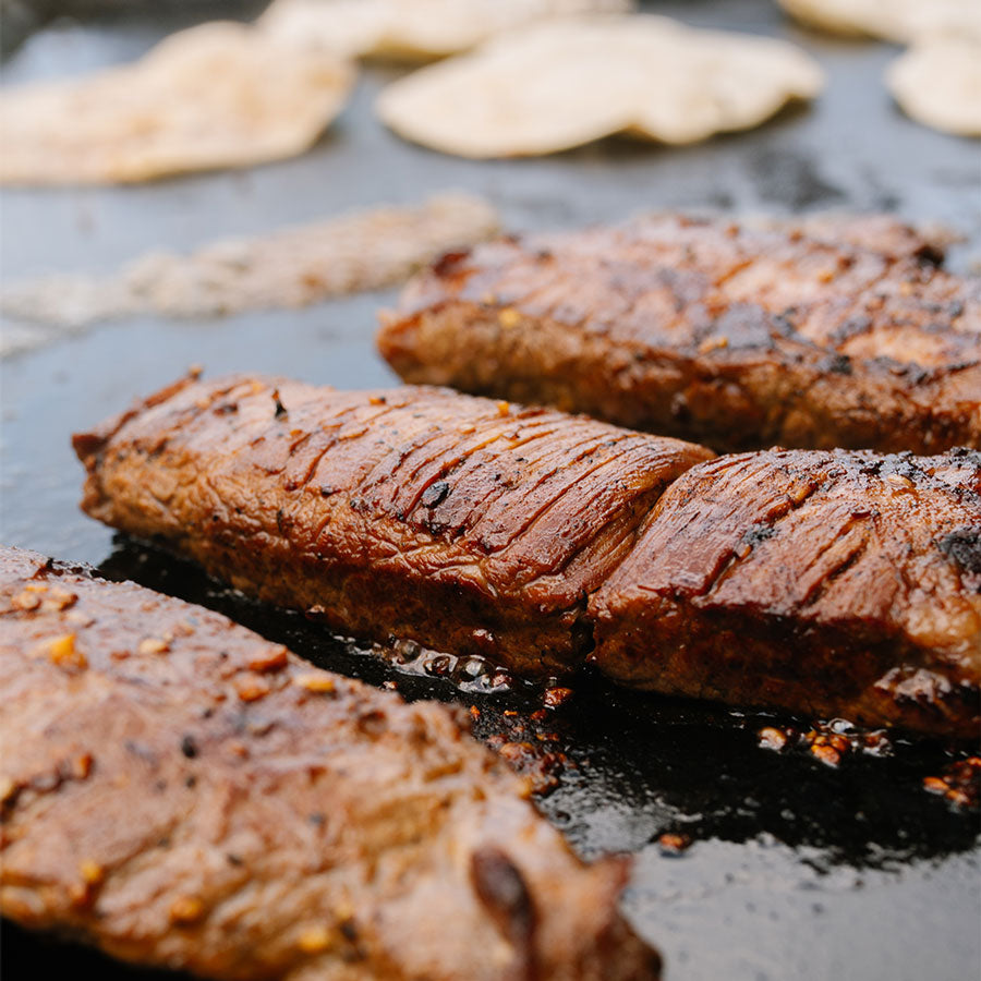Grilled steak strips with charred grill marks cooking on a dark skillet, with tortillas visible in the background.