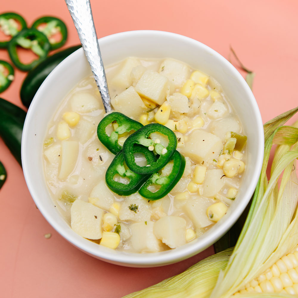 Beehive Meals Summer Corn Chowder topped with jalapeños in a white bowl on a pink background.