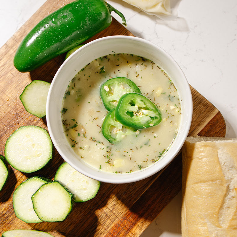 Bowl of Beehive Meals Summer Corn Chowder with jalapeño slices on a wooden cutting board with vegetables and cheese.