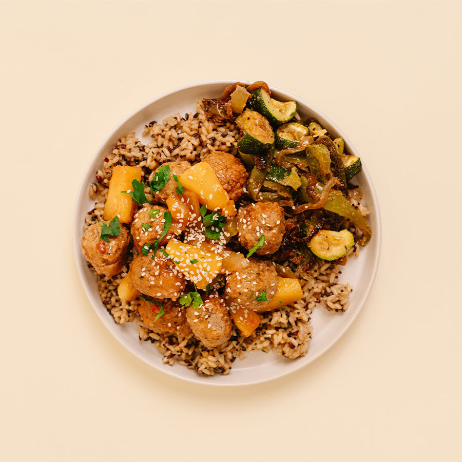 Overhead shot of a plated meal featuring glazed meatballs, sliced pineapple, zucchini, and brown rice on a light beige plate against a solid cream background.