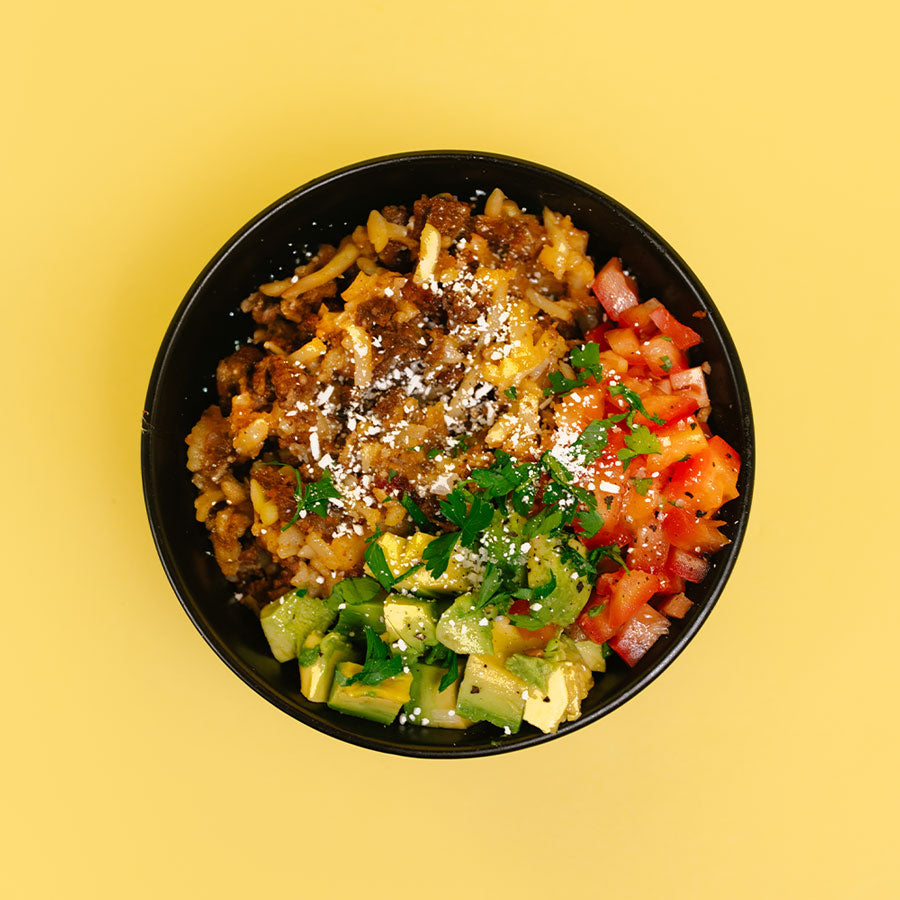 A top-down view of a black bowl filled with a colorful taco-style rice dish, topped with shredded cheese, white cheese flakes, diced red tomato, and chopped green cilantro, set against a solid yellow background.