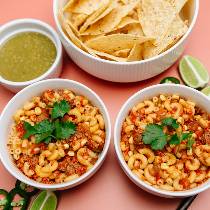 Two bowls of Beehive Meals Taco Mac with a side of tortilla chips and salsa on a pink background.